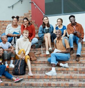 Full length of smiling young multi-ethnic male and female university students sitting on steps at community college